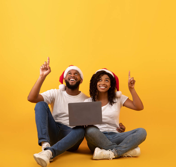 Cónyuges afroamericanos positivos en sombreros de Santa sentado con el ordenador portátil y apuntando hacia arriba en el espacio vacío sobre fondo amarillo - Foto, imagen