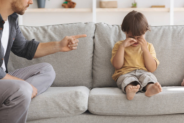 Lasten hyväksikäyttöä. Onneton pieni poika istuu sohvalla ja itkee, tunnistamaton vihainen isä torui järkyttynyt lapsi - Valokuva, kuva