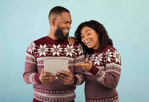 Emocionado casal preto em camisolas quentes usando tablet digital sobre fundo azul, fazendo compras on-line de Natal - Foto, Imagem