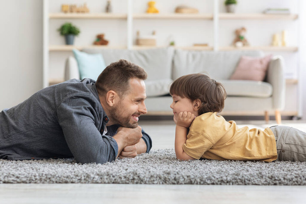 Zijaanzicht portret van knappe vader en zijn schattige zoontje kijken elkaar aan en glimlachen, liggend op de vloer thuis - Foto, afbeelding