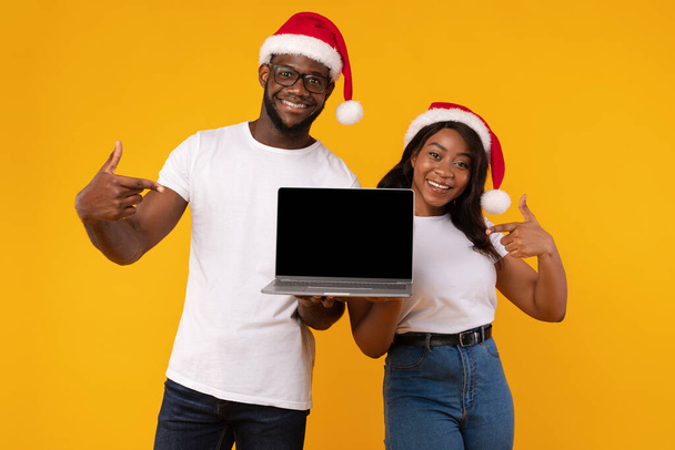Couple noir montrant l'écran d'ordinateur portable portant des chapeaux de Père Noël, fond jaune - Photo, image