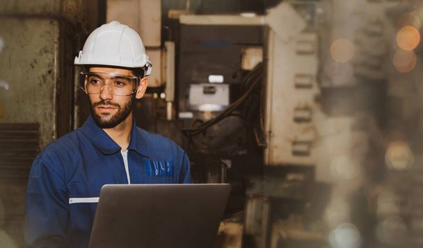Профессиональный красивый мужчина-инженер в синей форме и шлеме, работающий на ноутбуке в помещении механического завода. - Фото, изображение