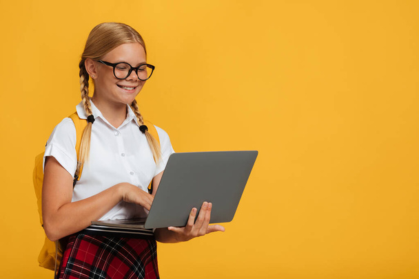 Χαρούμενη έξυπνη καυκάσια νεαρή μαθήτρια με γυαλιά που διαβάζει με φορητό υπολογιστή, απομονωμένη σε κίτρινο φόντο - Φωτογραφία, εικόνα