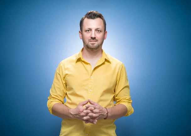 Serious young man standing with neutral face expression over blue background, dresses in yellow shirt. Bearded guy. Studio shot - Фото, изображение