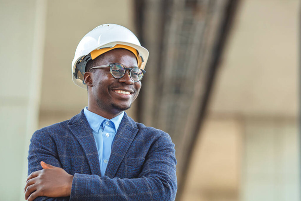 Portrait of african american man architect at building site with folded arms looking at camera. Confident construction manager in formal clothing wearing blue hardhat. Successful mature civil engineer at construction site with copy space. - Zdjęcie, obraz