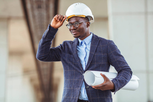 Portrait of multiethnic architect with blueprints. Mature African American contractor holding roll of architectural projects while looking at camera. Happy man in casual standing outdoor  - Photo, image