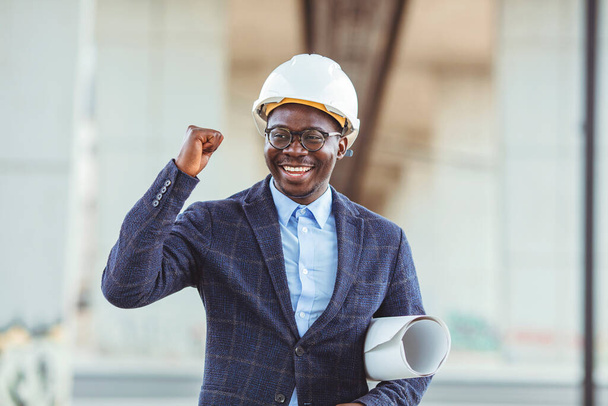 Happy smiling architect on a construction site with blueprints. Young architect man wearing contractor helmet holding project paper plan happy with big smile, excellent sign - Zdjęcie, obraz