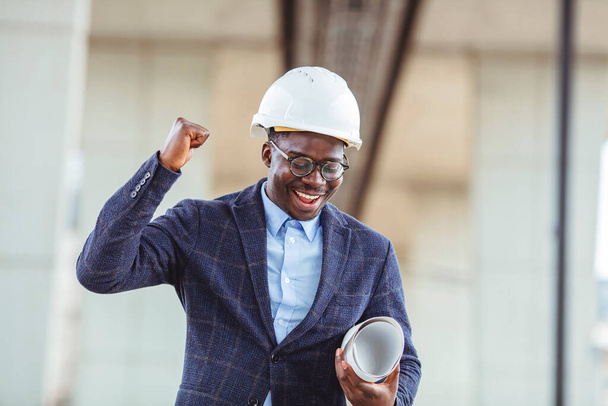 Content male construction architect in formal suit and hardhat with rolled blueprints, positive and happy expression. Young architect wearing a protective helmet standing on the building background - Foto, immagini
