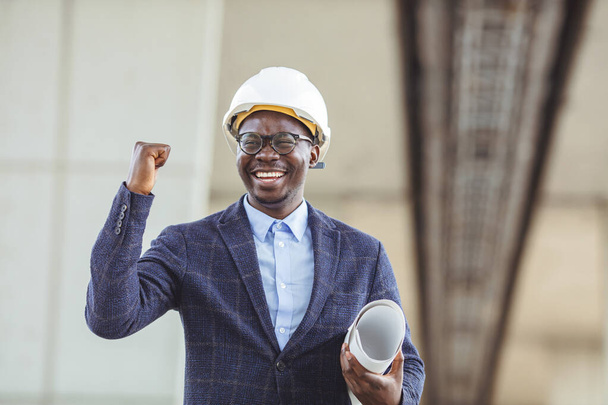 Content male construction architect in formal suit and hardhat with rolled blueprints, positive and happy expression. Young architect wearing a protective helmet standing on the building background - Foto, imagen