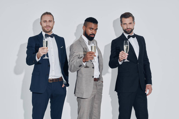 Three handsome young men in suits and bowties toasting with champagne while standing against gray background - Фото, изображение