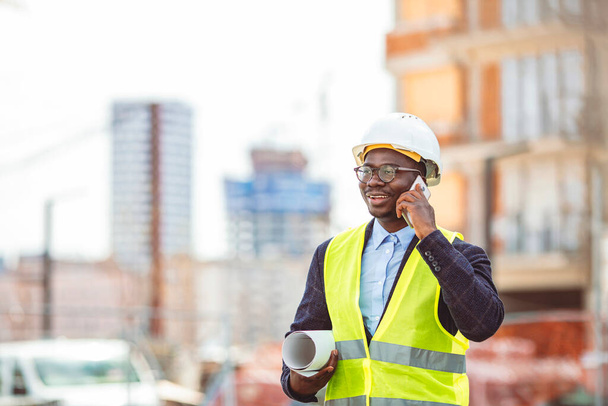 African American architect man holding blueprints using smartphone at street of city. Portrait of handsome young man architect on a building industry construction site - Foto, imagen