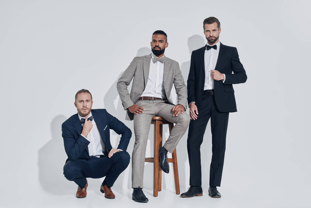 Three handsome young men in suits and bowties looking at camera while standing against gray background - Фото, изображение