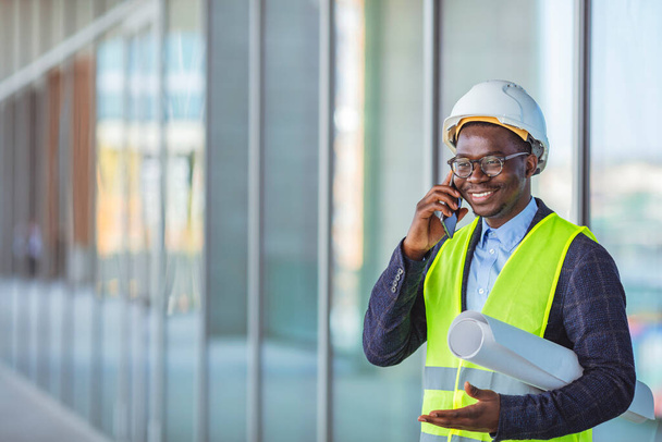 Smiling architect in suit and with helmet on head standing in building in construction process next to window and talking on smart phone. Engineer, architect or supervisor on a construction site of a new building, using a mobile app. - Foto, imagen