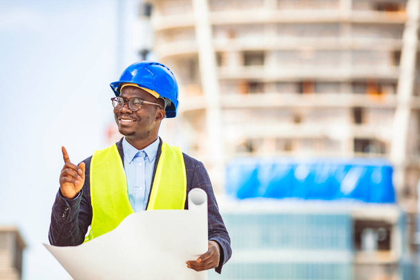 Portrait of attractive unshaven architect in suit with helmet on head walking around building in construction process and using tablet. Portrait of professional architect wearing blue helmet and looking at modern building outdoors - Photo, Image
