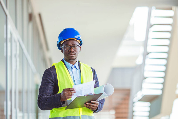Quality controller in factory. Portrait of smiling engineer in hardhat standing at construction site holding blueprint. Happy male architect writing on clipboard - Фото, изображение