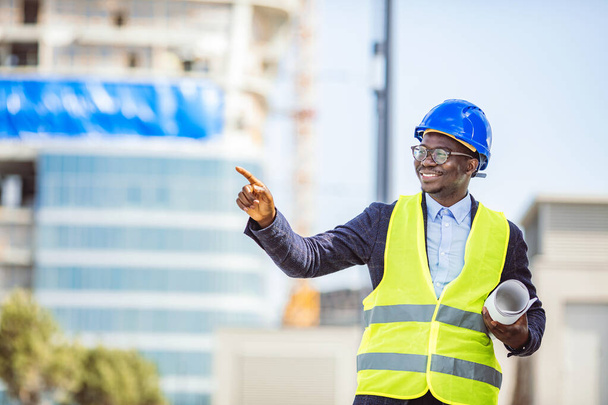 Portrait of smiling engineer in hardhat standing at construction site holding blueprint. Portrait Of happy young Construction Worker On Building Site. Professional engineer in safety equipment at construction site - Photo, Image