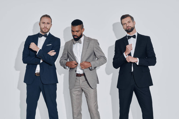 Three handsome young men in suits and bowties looking at camera while standing against gray background - Фото, изображение