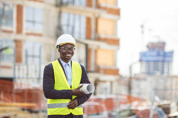 Portrait of African American man architect wearing a vest and helmet, he stands and holding a blueprints with the sky and urban in background. Engineer and architect concept. - Zdjęcie, obraz