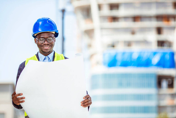 Engineer on site checking project. Portrait of smiling young architect or handsome engineer holding rolled up blueprints with building construction background. - 写真・画像