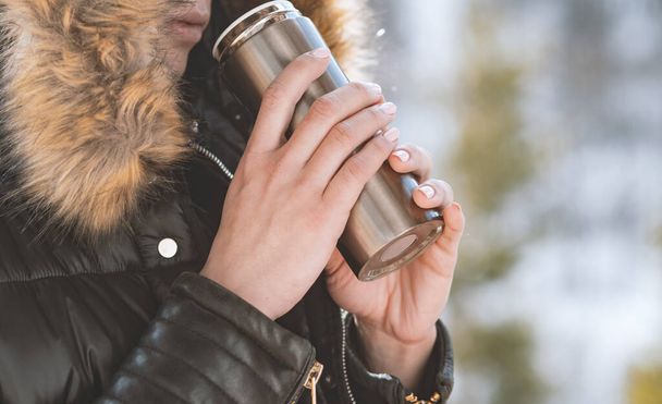 hot tea or coffee out of thermos in woman hands  on winter forest background. girl using a thermos in on a snowy mountain. - Photo, image