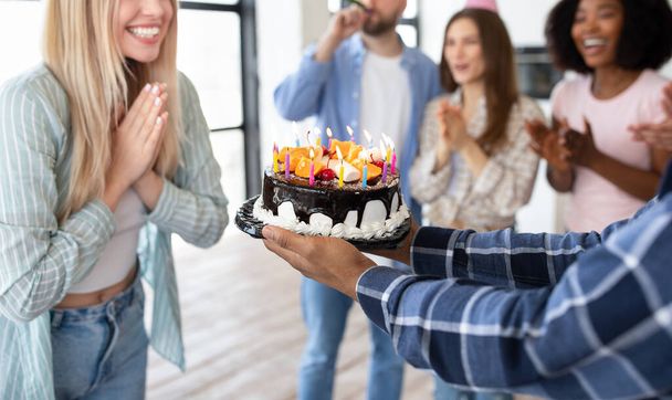 Vista de cerca de las personas alegres multirraciales que presentan pastel de cumpleaños con velas encendidas a la joven mujer rubia en casa - Foto, Imagen