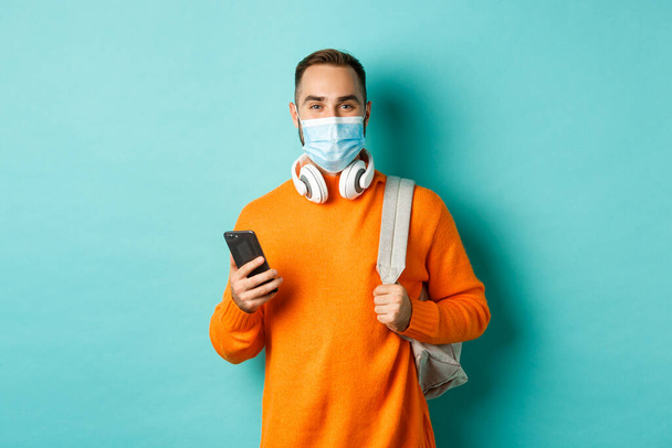 Young man in face mask using mobile phone, holding backpack, standing against light blue background - Photo, Image