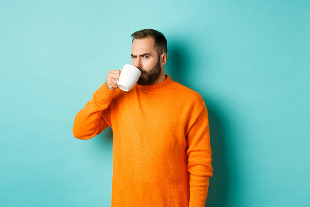 Imagem do cara bebendo café e olhando suspeito para a câmera, olhar para algo estranho, de pé em suéter laranja sobre fundo turquesa - Foto, Imagem