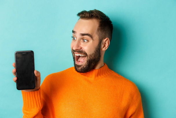 Close-up van de jonge man met de baard die een telefoonscherm toont en verbaasd kijkt, met oranje trui aan, staande tegen de achtergrond van de studio - Foto, afbeelding