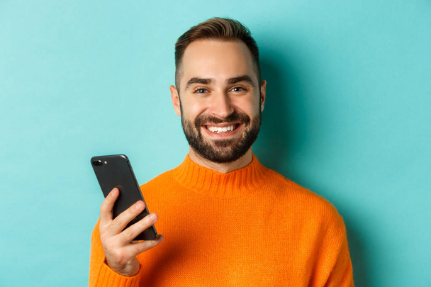 Primer plano del hombre guapo feliz escribiendo un mensaje en el teléfono móvil, sosteniendo el teléfono inteligente y sonriendo, de pie sobre fondo turquesa - Foto, imagen