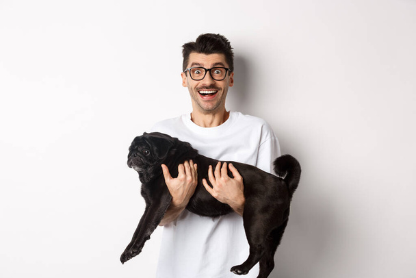 Cheerful handsome man carry black pug and smiling, staring at camera happy. Dog owner holding cute dog in arms, standing over white background - Foto, imagen