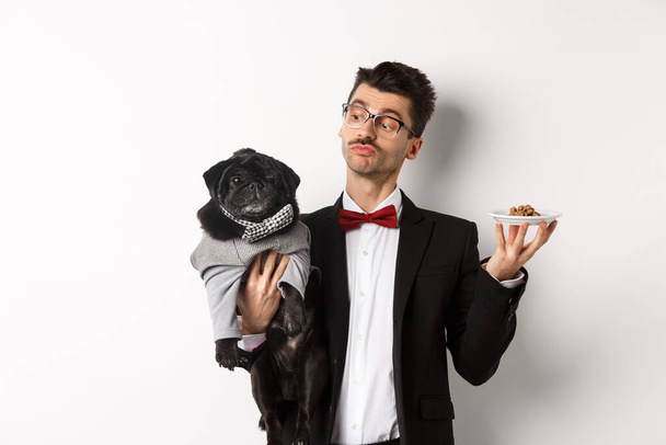 Hübsche junge Hundebesitzerin im schicken Anzug mit süßem schwarzen Mops und Teller mit Tierfutter vor weißem Hintergrund - Foto, Bild