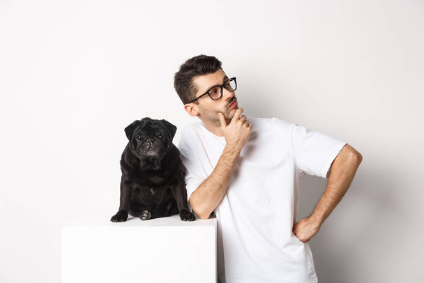 Image of handsome young man, pet owner standing with his dog and looking right at logo, posing with pug against white background - Foto, imagen