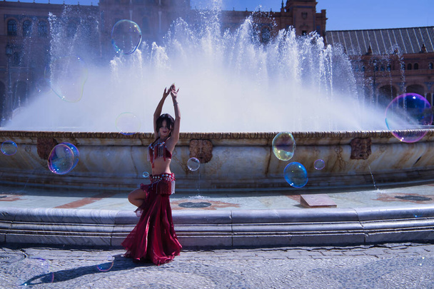 中世のヒスパニック系の女性は、ラインストーンと赤いドレスを着て、腹の踊りのために、噴水の前でシャボン玉に囲まれて踊る。ベリーダンスのコンセプト. - 写真・画像