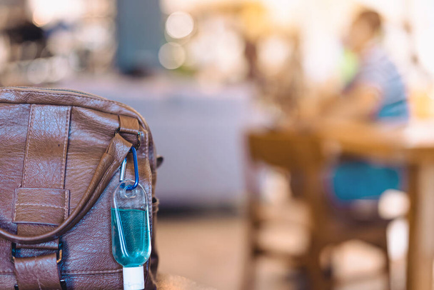 Corona Virüsü 'nü (Covid-19) öldürmek için mini portatif mavi alkol jeli şişesi kafeterya masasındaki bir kadının kahverengi deri çantasına asılır. Yeni normal yaşam tarzı. Alkol jeline odaklan. - Fotoğraf, Görsel