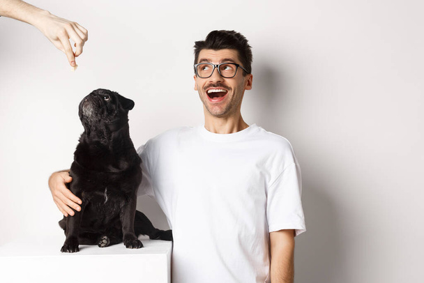 Verbazingwekkende jonge man in bril knuffelen zijn hond, huisdier eigenaar en pug staren naar de linker bovenhoek promo aanbod, staande over witte achtergrond - Foto, afbeelding