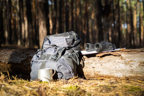 Tourist backpack, hat, binoculars and a map on a log in the forest. Concept hiking, hiking in the mountains. - Photo, Image