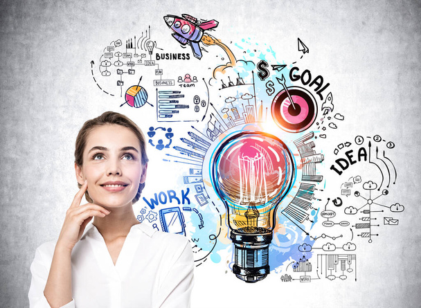 Smiling businesswoman in white blouse stands near sketch with light bulb, rocket launch, cogwheel, business, plan, teamwork on concrete wall. Concept of imagination and inspiration for creative ideas - Foto, immagini