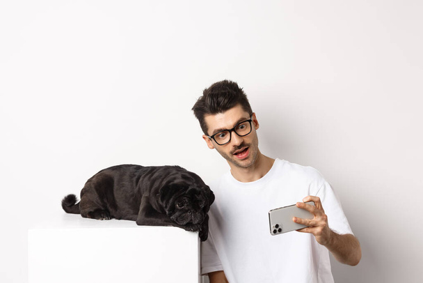 Hundebesitzer macht Selfie mit süßem Liegemops, macht Fotos mit Handy, weißer Hintergrund - Foto, Bild