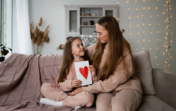 Η μαμά και η κόρη κάθονται στον καναπέ στο δωμάτιο και κρατούν μια κάρτα με καρδιά. - Φωτογραφία, εικόνα