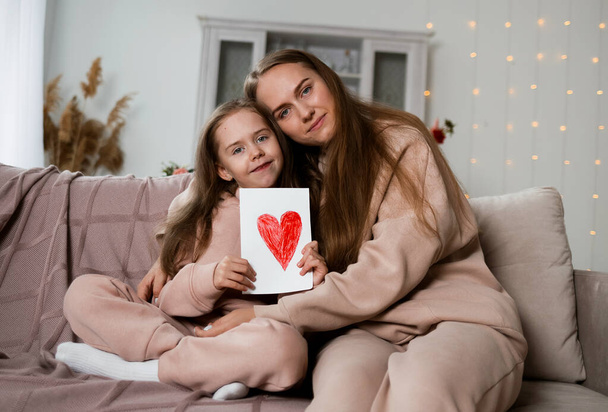 Мама и дочь сидят на диване в комнате с открыткой с сердцем и смотрят в камеру - Фото, изображение