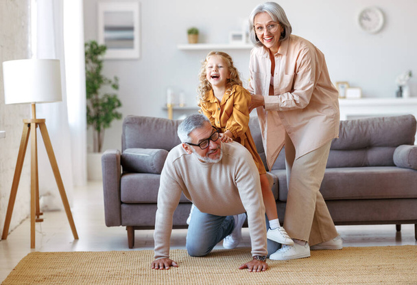 Aktywny pozytywny senior para dziadkowie bawiąc się z cute little girl wnuczka w salonie, zabawy i spędzania wolnego czasu w domu, uśmiechnięty dziadek niosąc podekscytowane dziecko na plecach - Zdjęcie, obraz