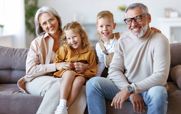 Boldog család örömteli kisgyerekek ölelkeznek pozitív idősebb nagyszülőkkel, miközben otthon ülnek a kanapén, vidám nagymama és nagypapa, a gyerekek mosolyognak a kamerába. - Fotó, kép