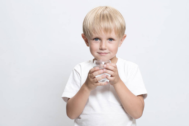 Un bel bambino caucasico, biondo con un bicchiere d'acqua. Sfondo da studio bianco. Concetto di acqua pulita che può essere bevuta dai bambini, problema mondiale delle risorse idriche e dell'acqua potabile. Copia spazio - Foto, immagini