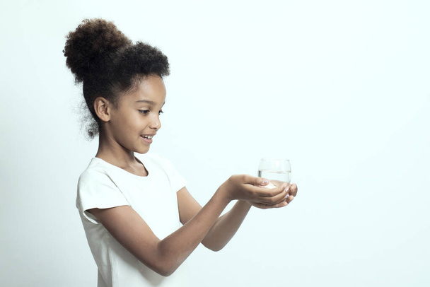 若い女の子は、プロファイルに立って、プレーン飲料水のガラスを保持します。ホワイトスタジオの背景。きれいな、精製された水、体のミネラルバランスの概念。水のガラスの子供。スペースのコピー. - 写真・画像