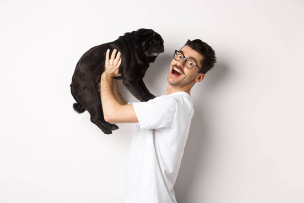 Wizerunek przystojnego młodzieńca kochającego swój mops. Właściciel psa trzyma szczeniaka i uśmiecha się szczęśliwy przed kamerą, stojąc na białym tle - Zdjęcie, obraz