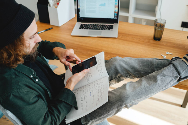 Bearded student zit met zijn voeten op zijn bureau terwijl hij online studeert met behulp van laptop en smartphone. College student voor te bereiden op universitaire les - Foto, afbeelding