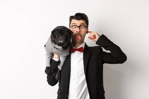Забавный молодой человек в костюме для вечеринки, показывающий язык и держащий милого черного мопса на плече, празднующий с домашним животным, стоящий на белом фоне - Фото, изображение