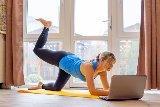 Beautiful young woman in sportswear doing sport exercises on yoga mat at home. Fitness training online with a laptop. Healthy lifestyle concept. - Foto, Bild