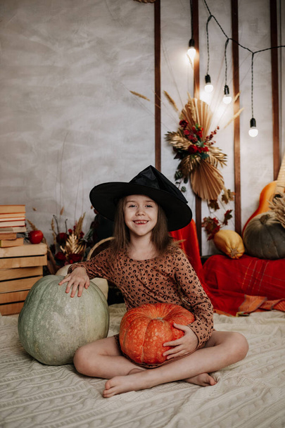 Cadı şapkası takmış küçük mutlu bir kız örgü örmüş bir battaniyenin üzerinde balkabağıyla oturuyor. - Fotoğraf, Görsel