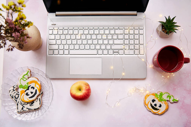 Gezellig thuis kantoor in Halloween: laptop en rode beker, zelfgemaakte koekjes in de vorm van schattige pompoenen. Atmosferische werksfeer met bloemenslingers. Kopieerruimte - Foto, afbeelding